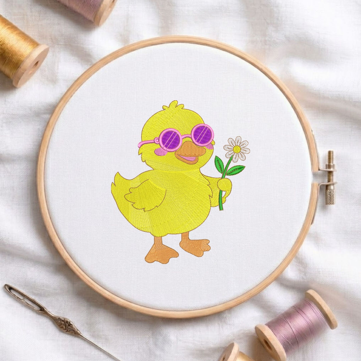 A yellow embroidered duckling wearing pink sunglasses and holding a white flower, stitched inside a wooden embroidery hoop on white fabric, surrounded by spools of thread and a needle.