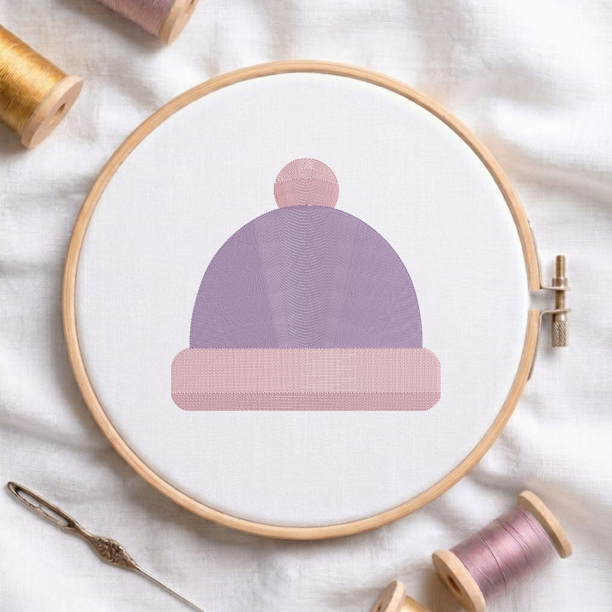 A circular embroidery hoop holding white fabric with a stylized knit-beanie design in lavender and dusty pink thread, styled on a white fabric background with spools of thread and a needle.