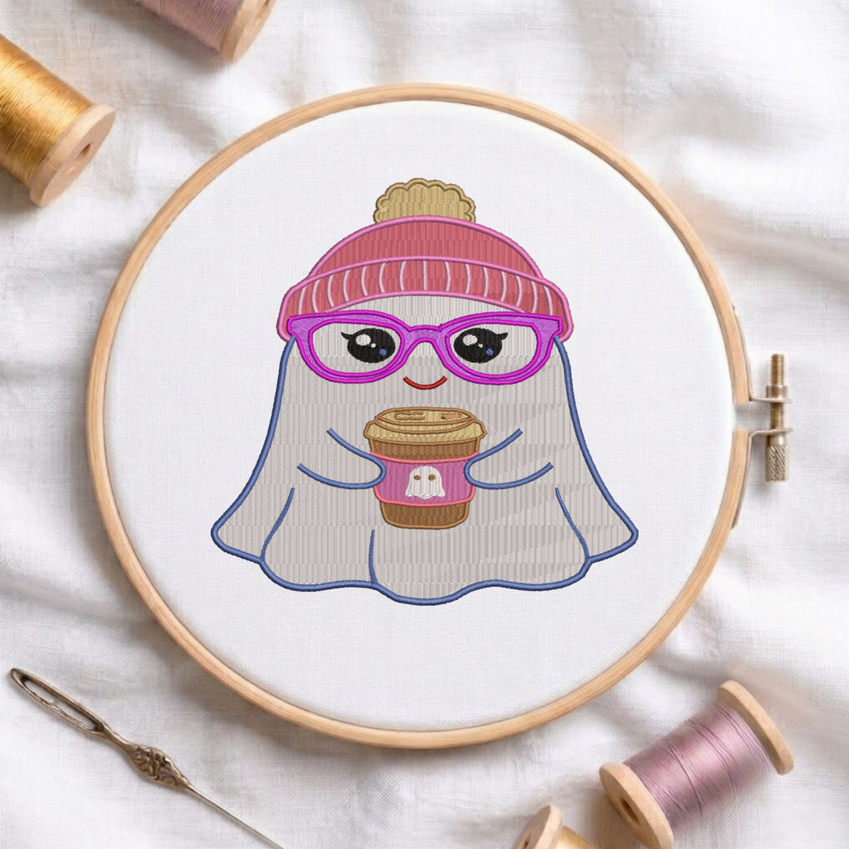 A circular embroidery hoop holding a white fabric design of a cute ghost character wearing a pink beanie and purple glasses, holding a coffee cup. The hoop is styled on a white fabric background with spools of thread and a needle.