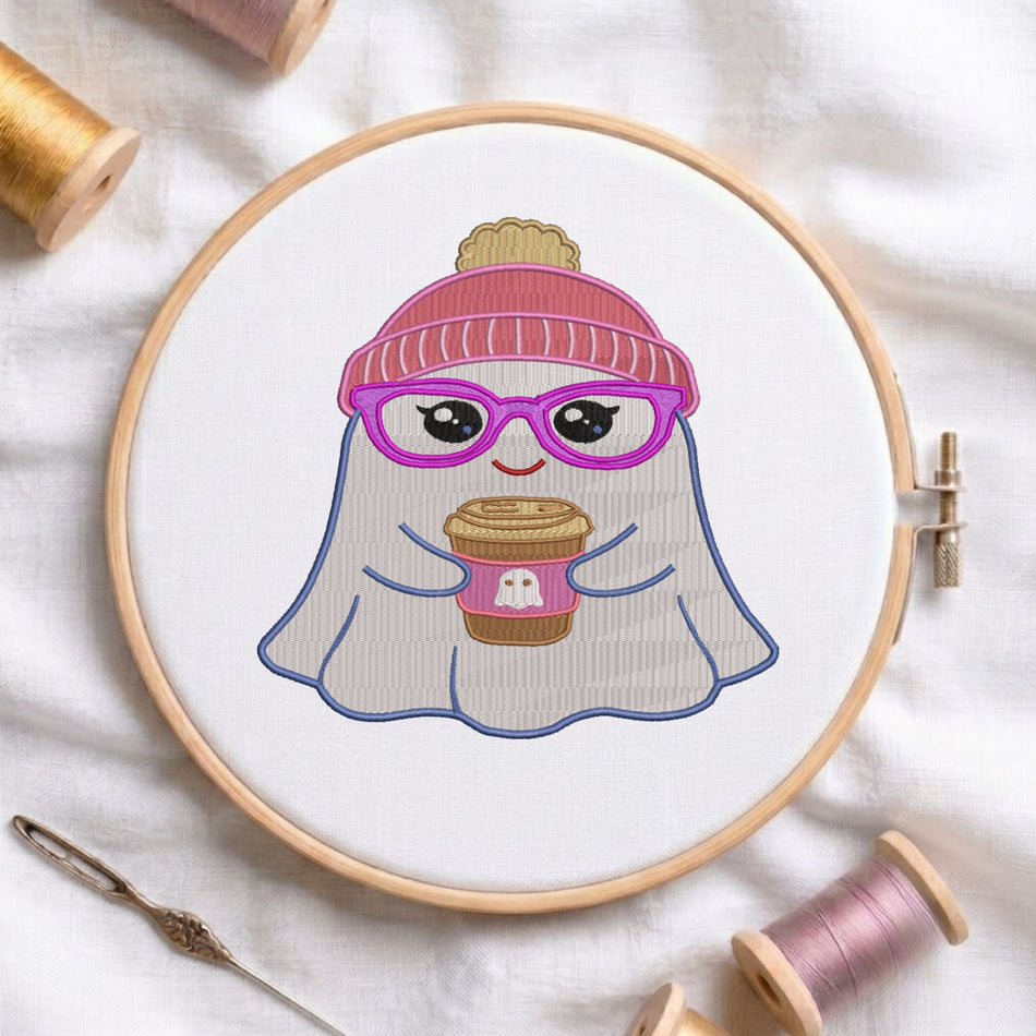 A circular embroidery hoop holding a white fabric design of a cute ghost character wearing a pink beanie and purple glasses, holding a coffee cup. The hoop is styled on a white fabric background with spools of thread and a needle.