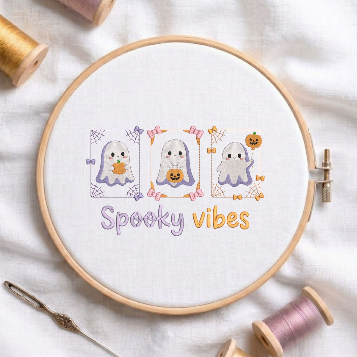 Cute spooky vibes ghost with pumpkins machine embroidery design displayed in a wooden hoop on white fabric with thread spools and a needle