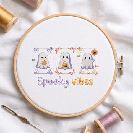 Cute spooky vibes ghost with pumpkins machine embroidery design displayed in a wooden hoop on white fabric with thread spools and a needle