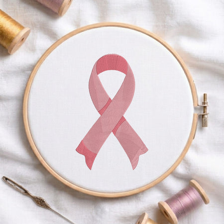 A pink awareness ribbon design stitched or printed on white fabric inside a wooden embroidery hoop, surrounded by spools of thread and a needle on a white fabric background.