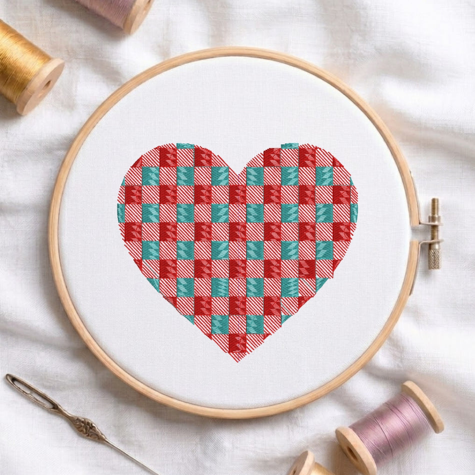 A heart-shaped cross-stitch design in a red and teal gingham pattern, held in a wooden embroidery hoop on a white fabric background, surrounded by spools of thread and a needle.