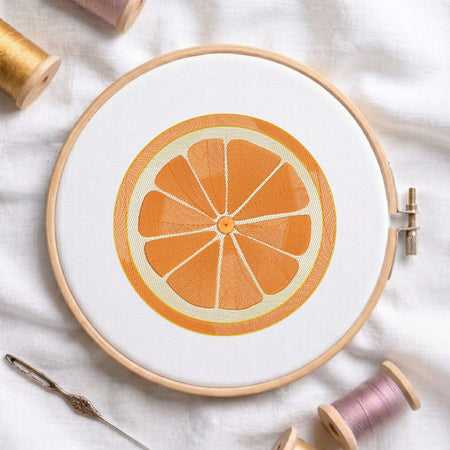 A circular embroidery hoop holding white fabric with an orange slice design stitched in orange and cream thread, styled on a white fabric background with spools of thread and a needle nearby.
