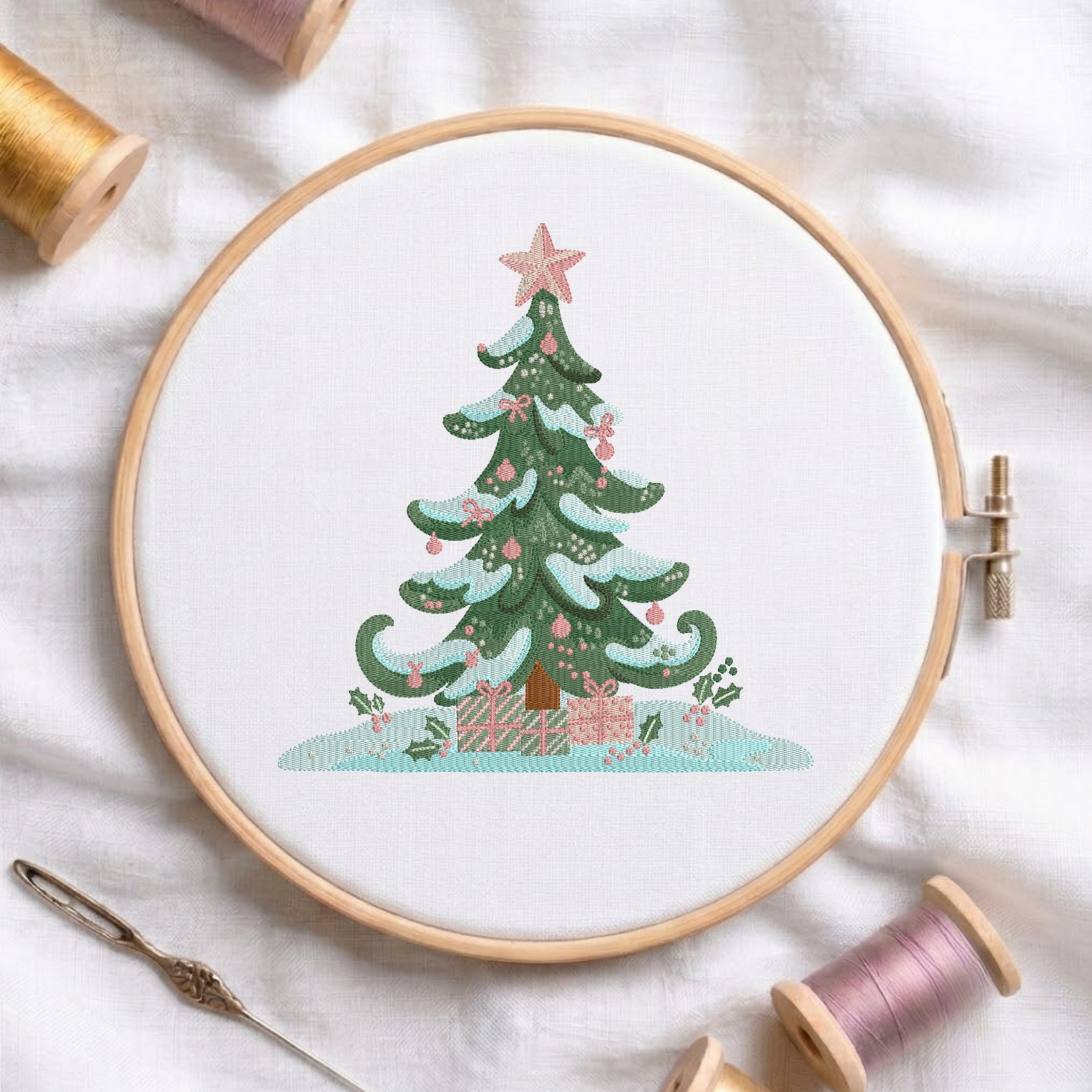 A circular embroidery hoop on white fabric, featuring a stitched Christmas tree with pink ornaments and gifts, surrounded by spools of thread and a needle as styled props.