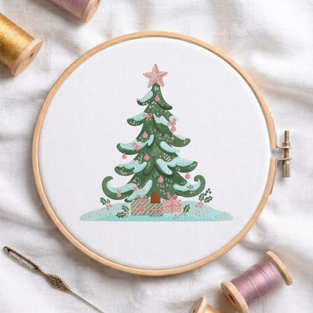 A circular embroidery hoop on white fabric, featuring a stitched Christmas tree with pink ornaments and gifts, surrounded by spools of thread and a needle as styled props.