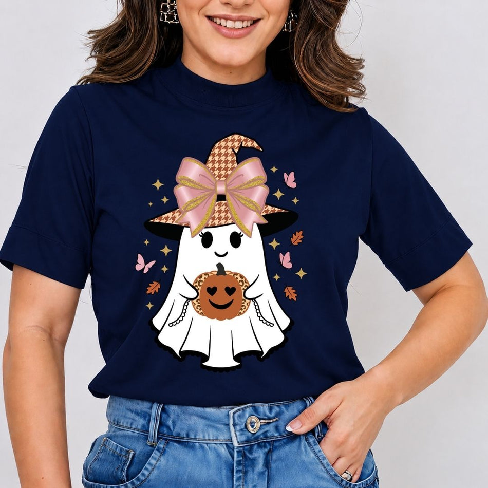A woman wearing a navy blue short-sleeve t-shirt with a cute Halloween graphic of a white ghost wearing a pink bow and houndstooth witch hat, holding a smiling pumpkin. She is paired with blue jeans against a light gray background.
