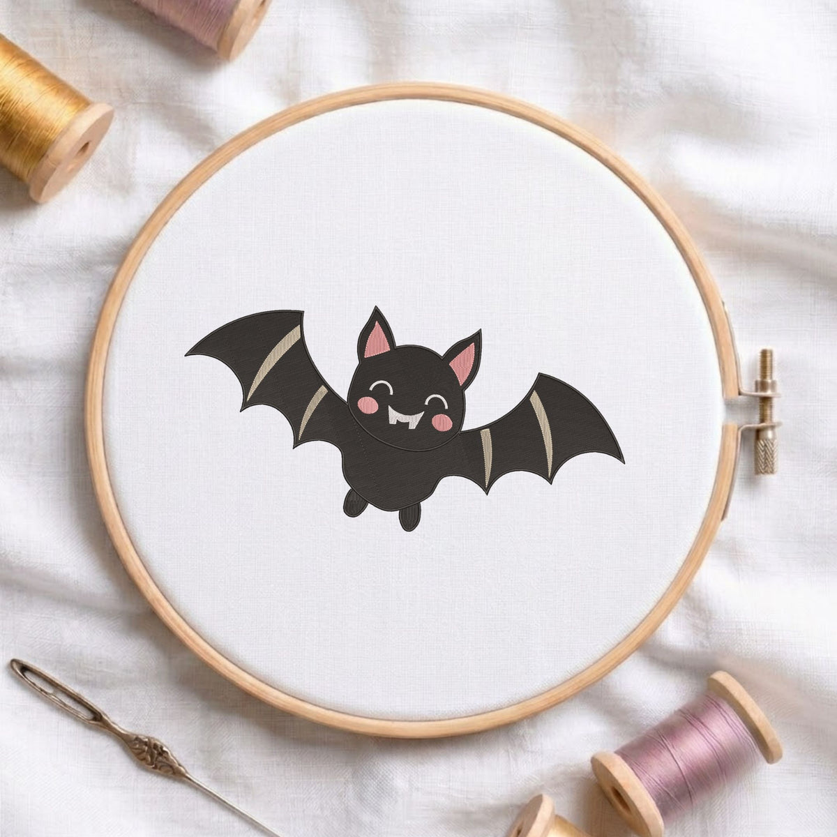 Cute flying Halloween bat machine embroidery design with pink ears, rosy cheeks, and little fangs, displayed inside a round wooden hoop on white linen fabric alongside thread spools and a needle.