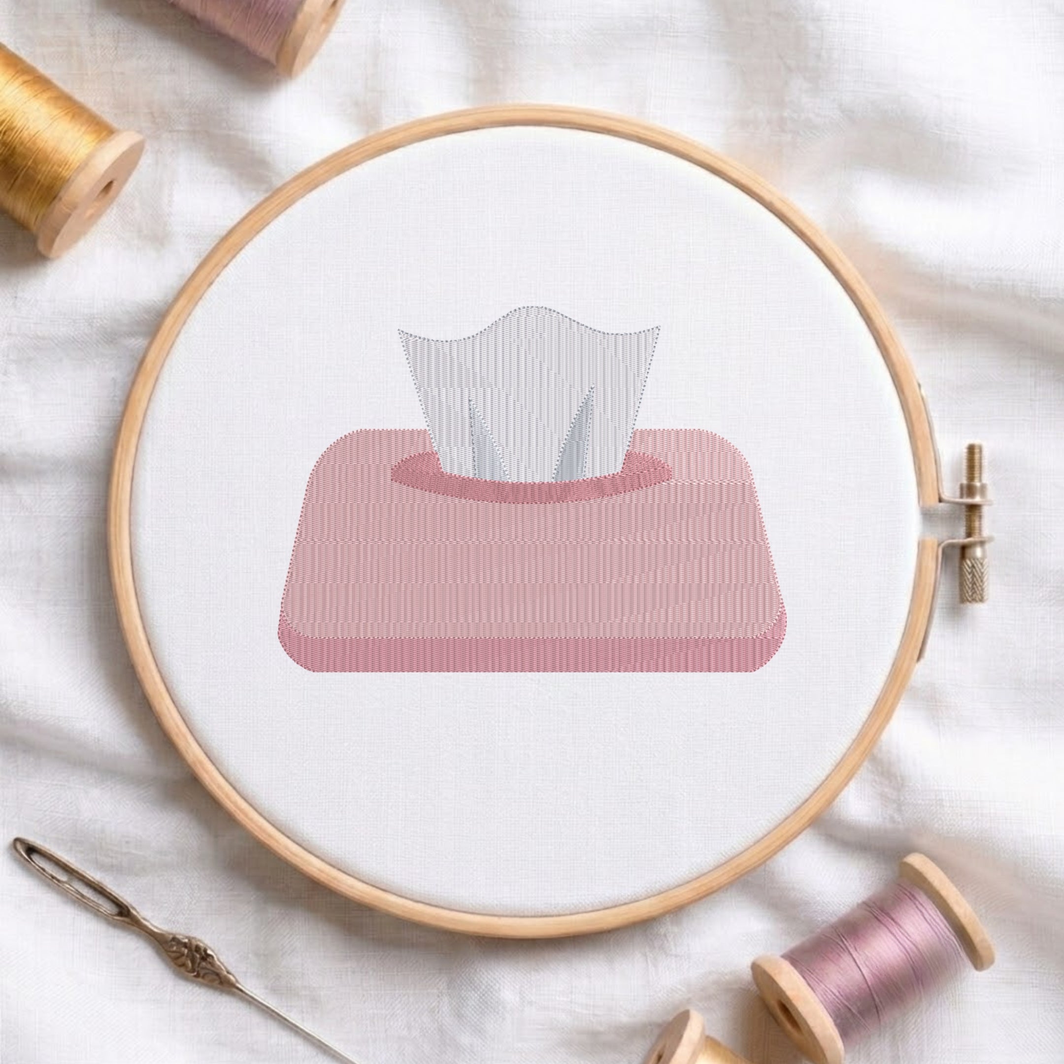 An embroidered tissue box design with a pink container and white tissues pulling out, stitched inside a wooden hoop on white fabric with sewing threads.