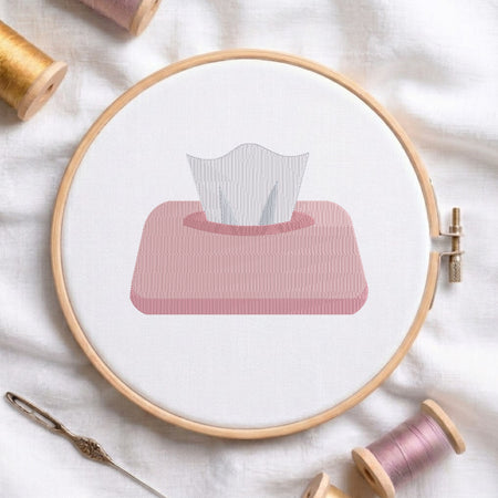 An embroidered tissue box design with a pink container and white tissues pulling out, stitched inside a wooden hoop on white fabric with sewing threads.