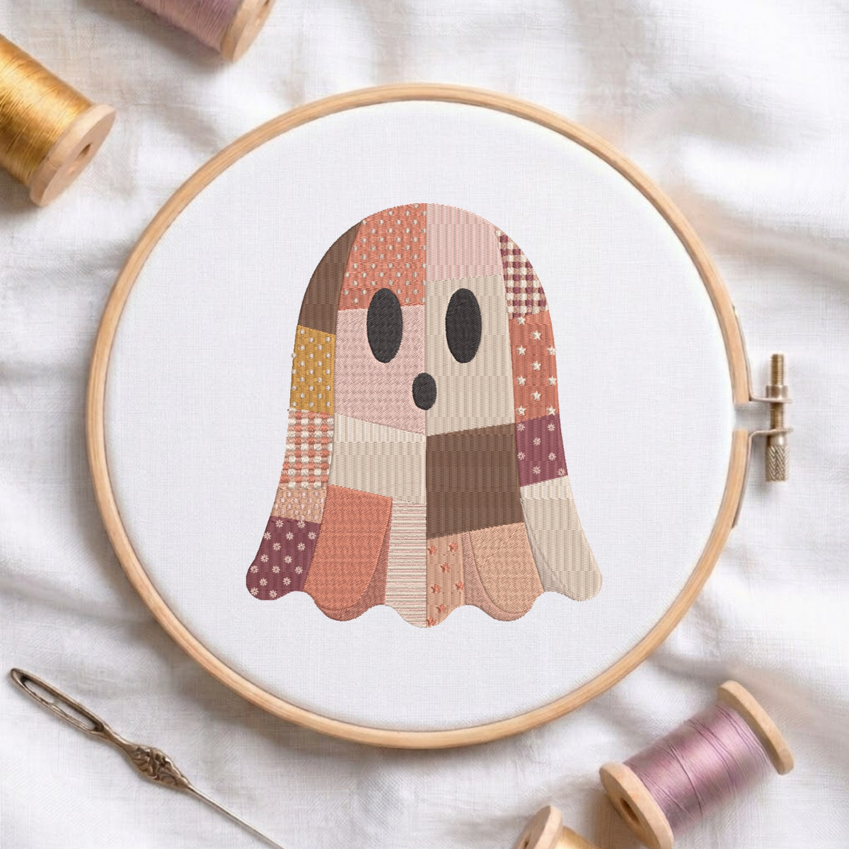 Cute patchwork Halloween ghost machine embroidery design displayed inside a round wooden hoop on white linen fabric, accompanied by wooden thread spools and a needle.