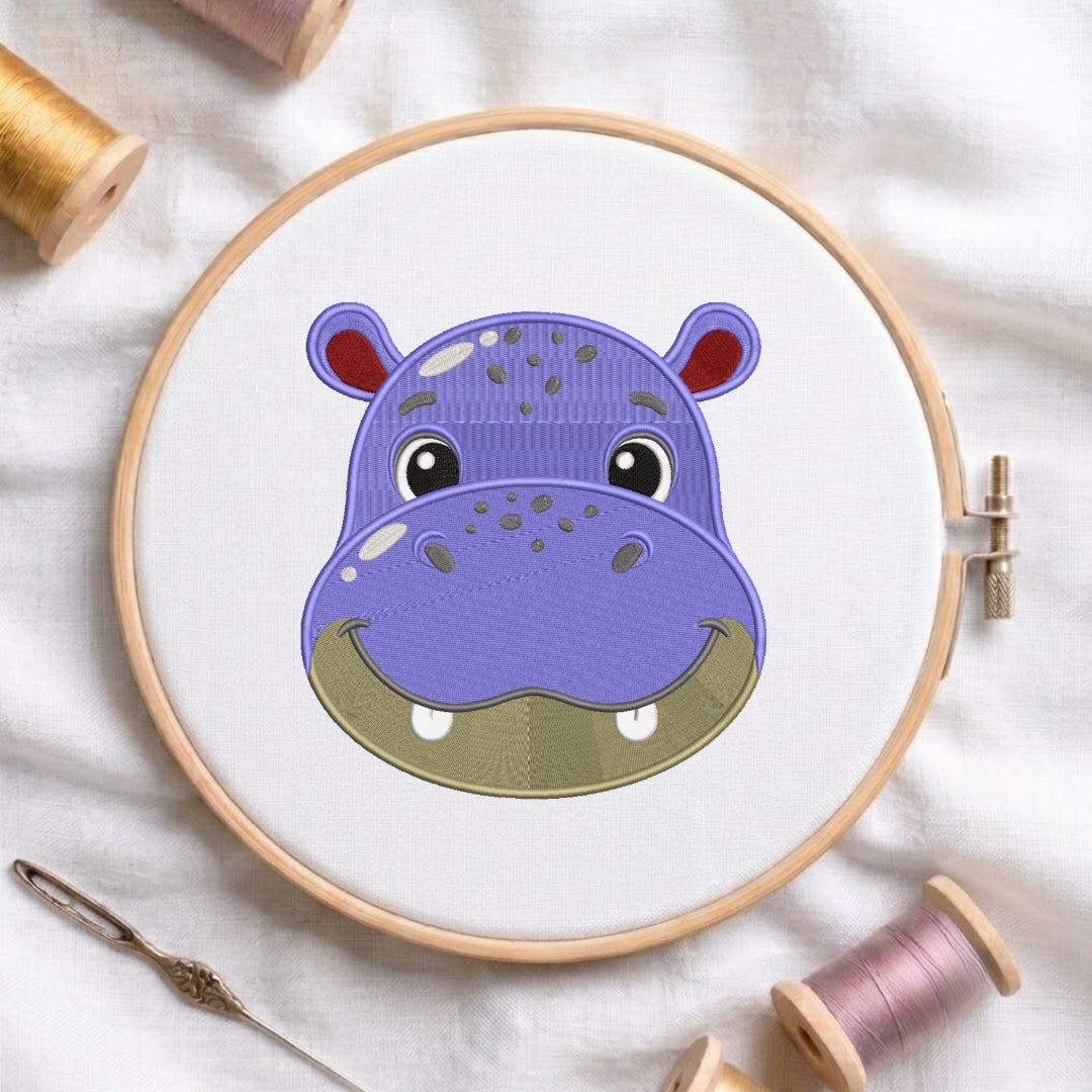 A circular embroidery hoop holding white fabric with a purple hippo face design, surrounded by spools of thread and a needle on a white fabric background.
