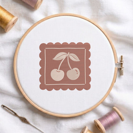 Vintage double cherry machine embroidery design framed inside a scalloped square border, displayed in a wooden hoop on white fabric with thread spools and a needle.