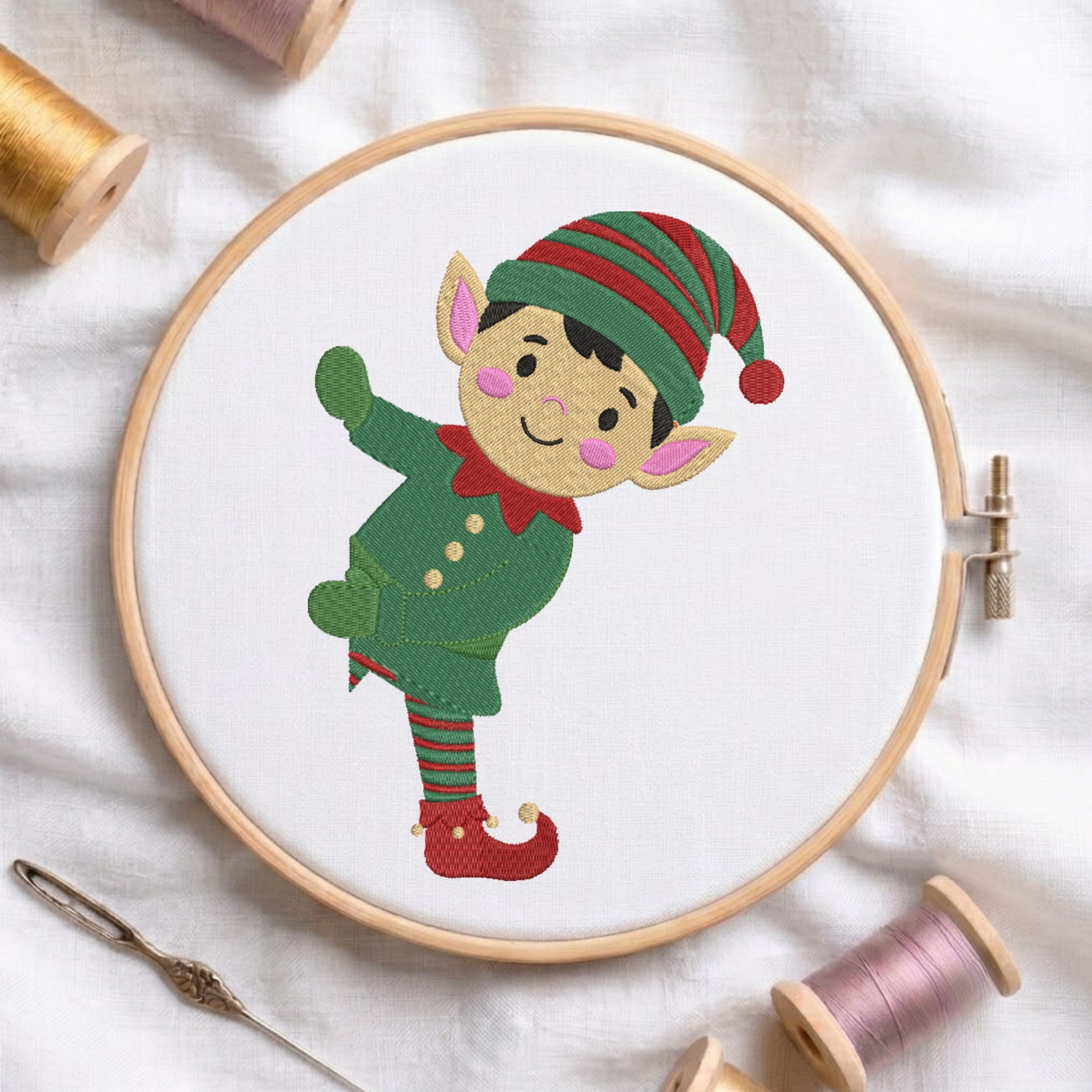 A circular embroidery hoop holding white fabric with a stitched elf character in green and red, surrounded by spools of thread and a needle on a white fabric background.