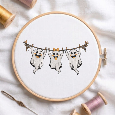 A circular embroidery hoop holding white fabric with a stitched design of three hanging ghosts on a clothesline, surrounded by spools of thread and a needle on a white fabric background.