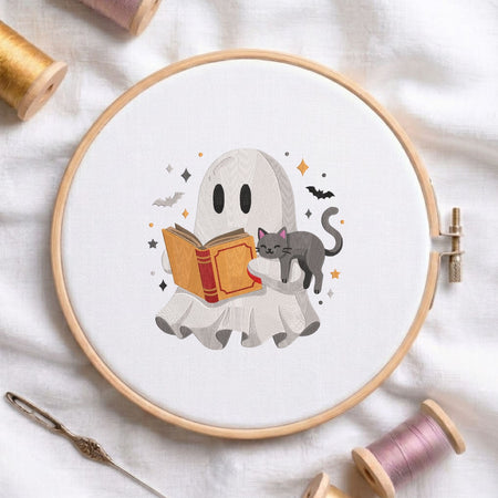 Cute ghost reading a book with a sleeping black cat machine embroidery design displayed in a wooden hoop on white fabric with thread spools and a needle.