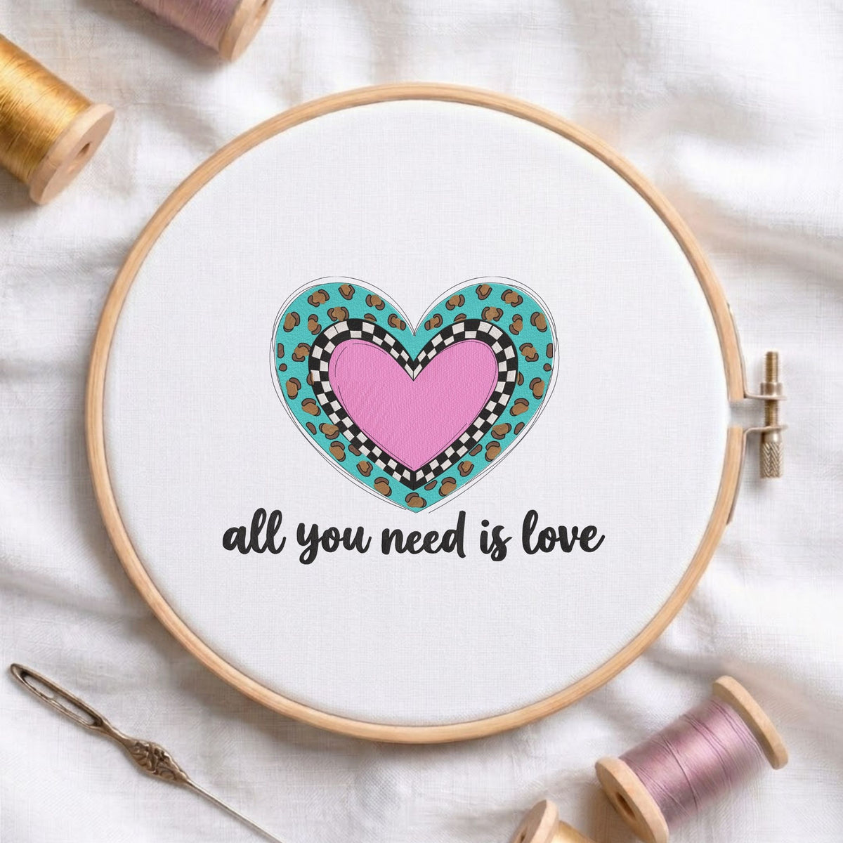 All you need is love" phrase with a layered pink heart, black and white checkerboard pattern, and turquoise leopard print machine embroidery design in a wooden embroidery hoop on white fabric with thread spools and a needle