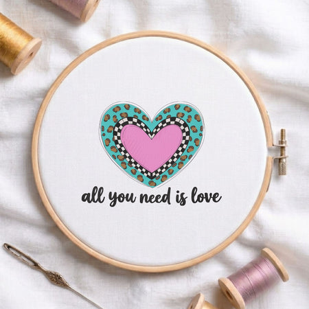 All you need is love" phrase with a layered pink heart, black and white checkerboard pattern, and turquoise leopard print machine embroidery design in a wooden embroidery hoop on white fabric with thread spools and a needle