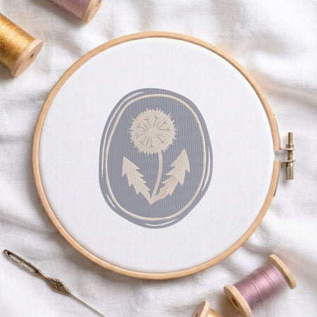 Vintage botanical dandelion flower in an oval frame machine embroidery design, displayed inside a round wooden hoop on white fabric with thread spools and a needle.
