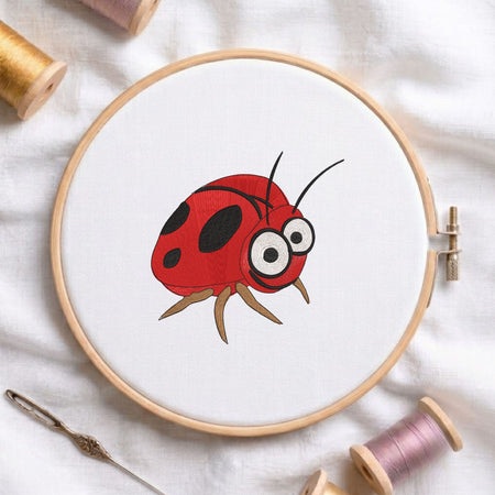 Cute funny ladybug machine embroidery design displayed in a wooden hoop on white fabric with thread spools and a needle