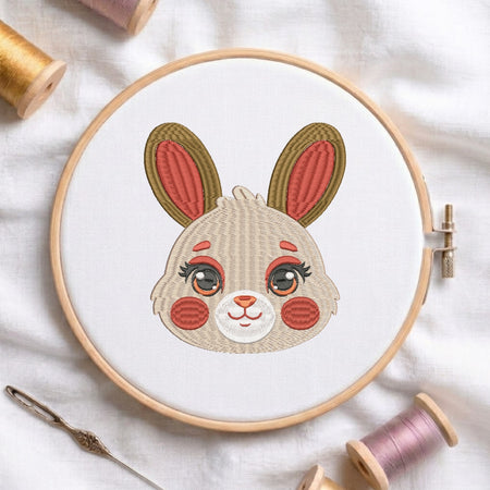 A circular embroidery hoop holding white fabric with a stylized bunny face design in beige, pink, olive green, and brown thread. The hoop is photographed on a white fabric surface with spools of thread and a needle around it.