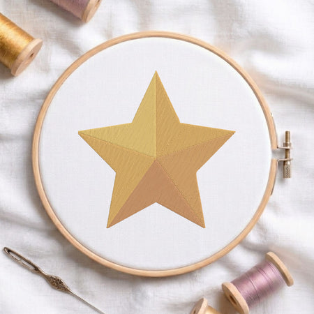 A circular embroidery hoop holding white fabric with a stitched gold star design, surrounded by spools of thread and a needle on a soft white fabric background.