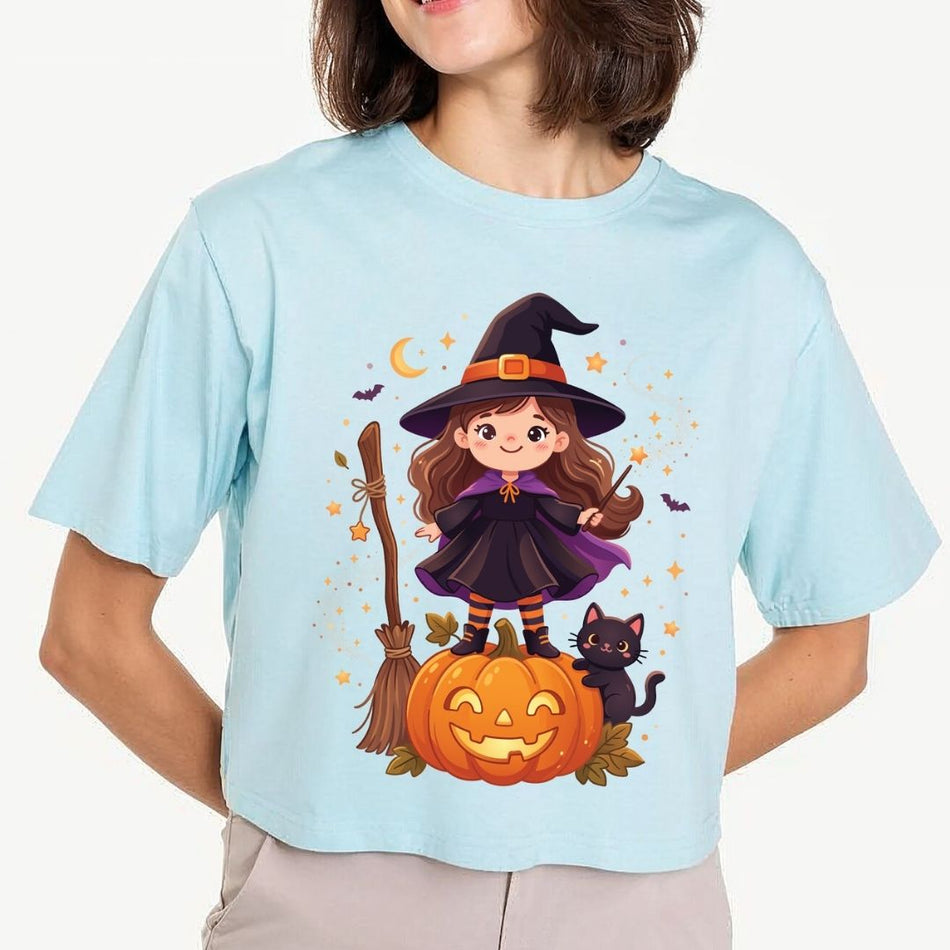 A woman wearing a light baby blue cropped t-shirt featuring a cartoon illustration of a cute little witch standing on a jack-o'-lantern pumpkin with a black cat.