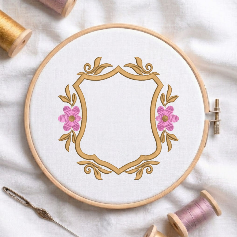 A circular embroidery hoop holding white fabric with a decorative gold crest and pink flowers, surrounded by spools of thread and a needle on a white fabric background.