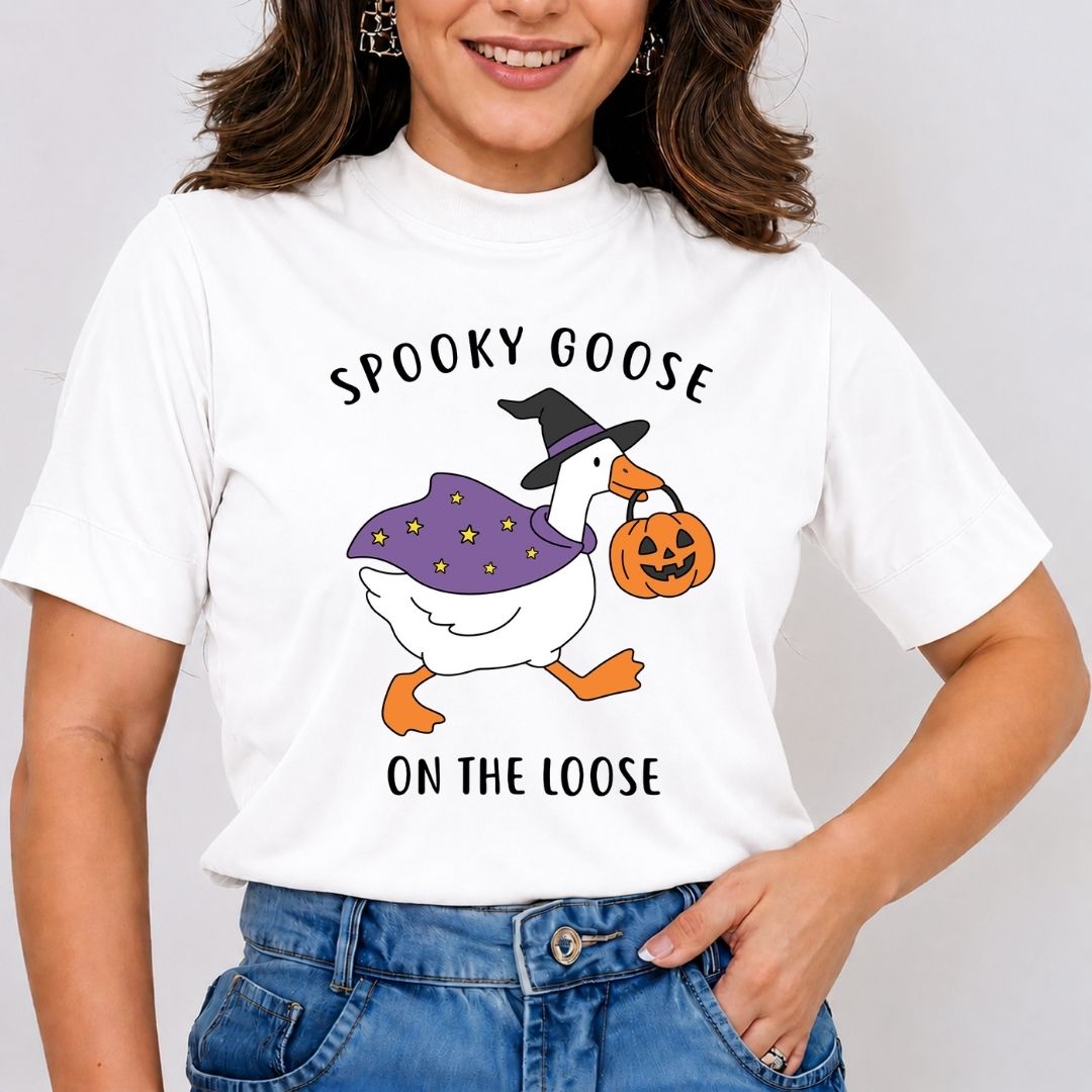 A person wearing a white t-shirt featuring an illustration of a goose wearing a witch hat and starry cape while carrying a jack-o-lantern bucket with the text Spooky Goose on the Loose.
