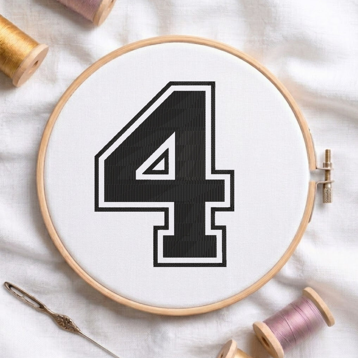 A wooden embroidery hoop holding white fabric with a large black number 4 outlined in white, styled on a white fabric background with spools of thread and a needle.
