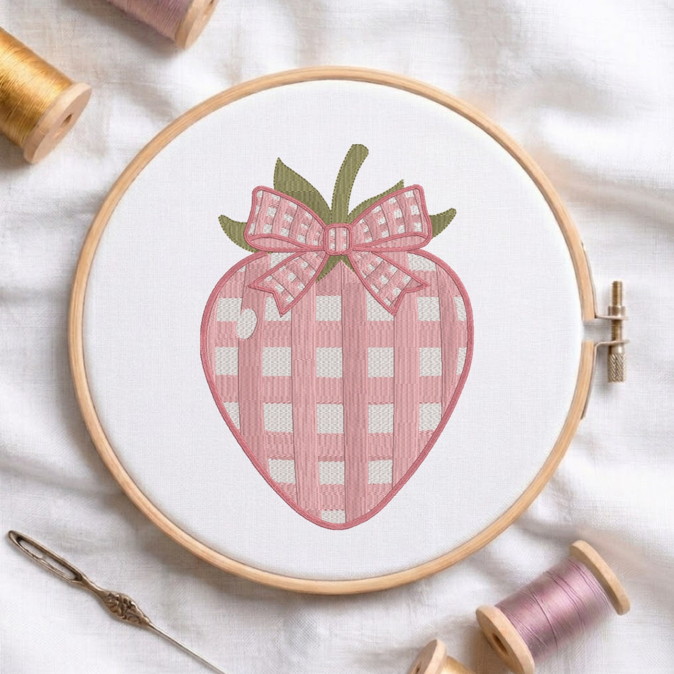 A circular embroidery hoop holding white fabric with a stitched strawberry design in pink gingham and green leaves, surrounded by spools of thread and a needle on a white fabric background.