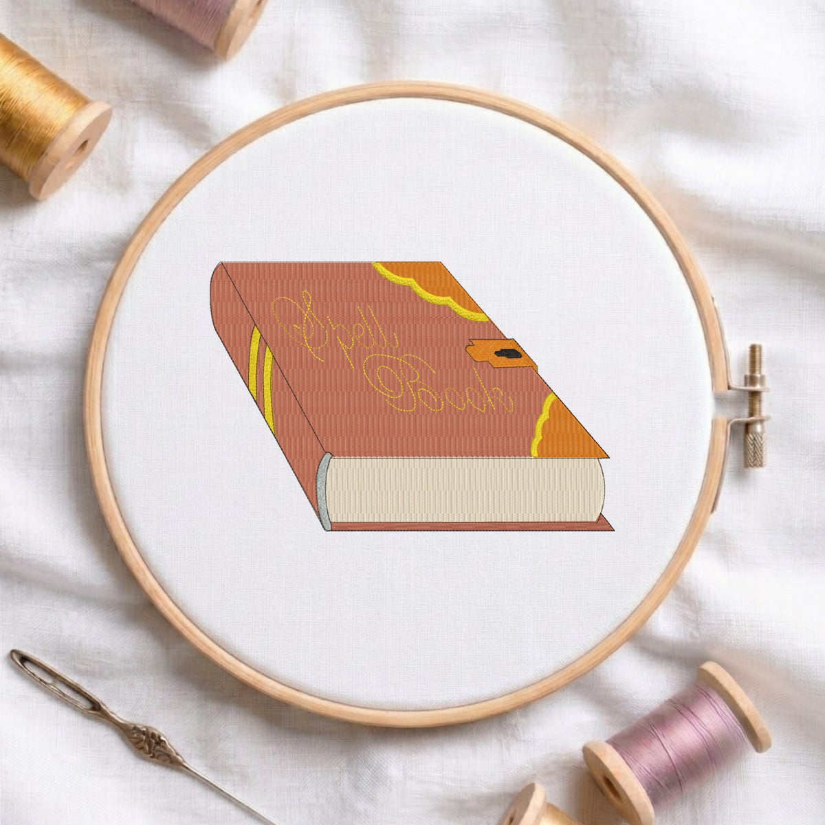 Vintage spell book with yellow cursive text and lock detail machine embroidery design displayed inside a round wooden hoop on white linen fabric, accompanied by wooden thread spools and a needle.