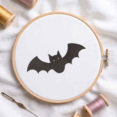 Spooky cute black bat machine embroidery design displayed inside a round wooden hoop on white fabric alongside thread spools and a needle.