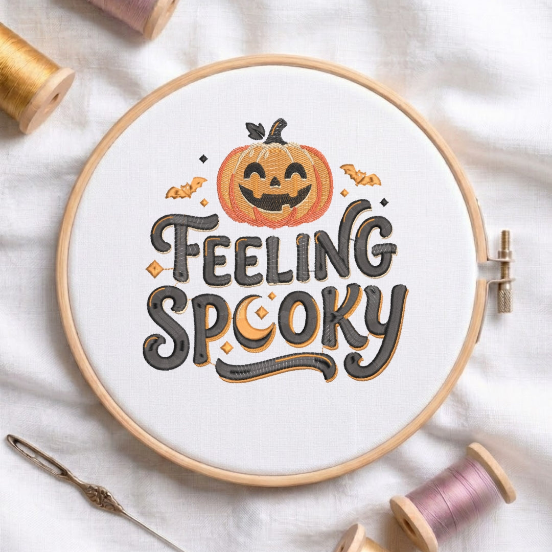 A circular embroidery hoop on white fabric, featuring a Halloween-themed design with a smiling orange pumpkin and the words “FEELING SPOOKY” in dark gray lettering with orange and black accents. Spools of thread and a needle are arranged around it on a white fabric background.