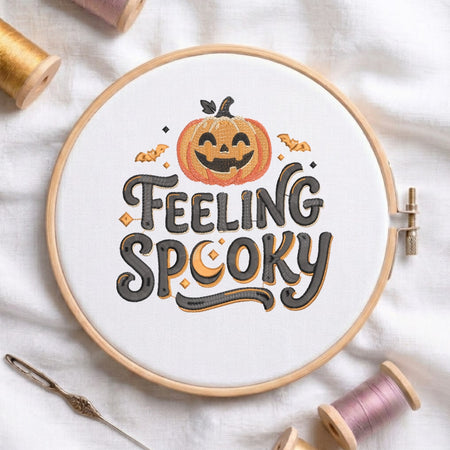 A circular embroidery hoop on white fabric, featuring a Halloween-themed design with a smiling orange pumpkin and the words “FEELING SPOOKY” in dark gray lettering with orange and black accents. Spools of thread and a needle are arranged around it on a white fabric background.