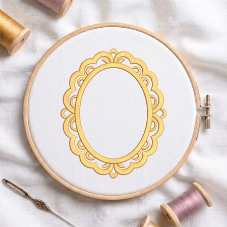 A circular embroidery hoop holding white fabric with a decorative gold ornamental frame design, styled on a white cloth with thread spools and a needle around it.