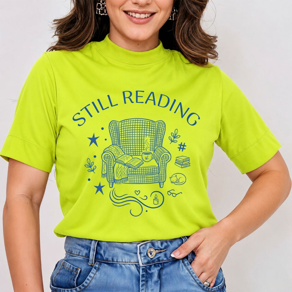 Woman wearing a bright lime green mock neck t-shirt featuring a blue retro line art "Still Reading" graphic print with a cozy armchair, book stack, and sleeping cat, tucked into jeans.