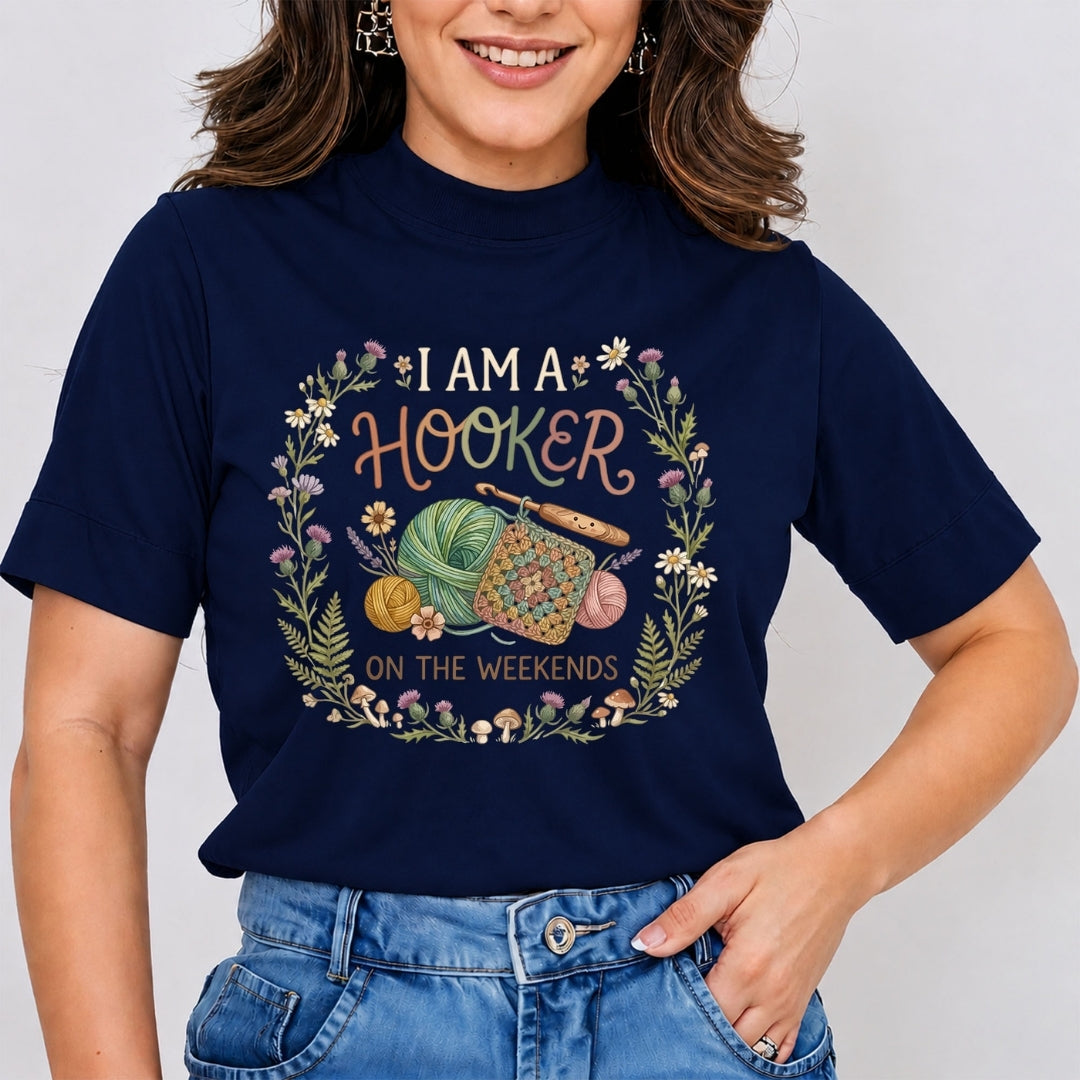 A woman wearing a navy blue graphic t-shirt with a floral wreath design and the text 'I AM A HOOKER ON THE WEEKENDS' along with knitting needles and yarn, paired with blue jeans.