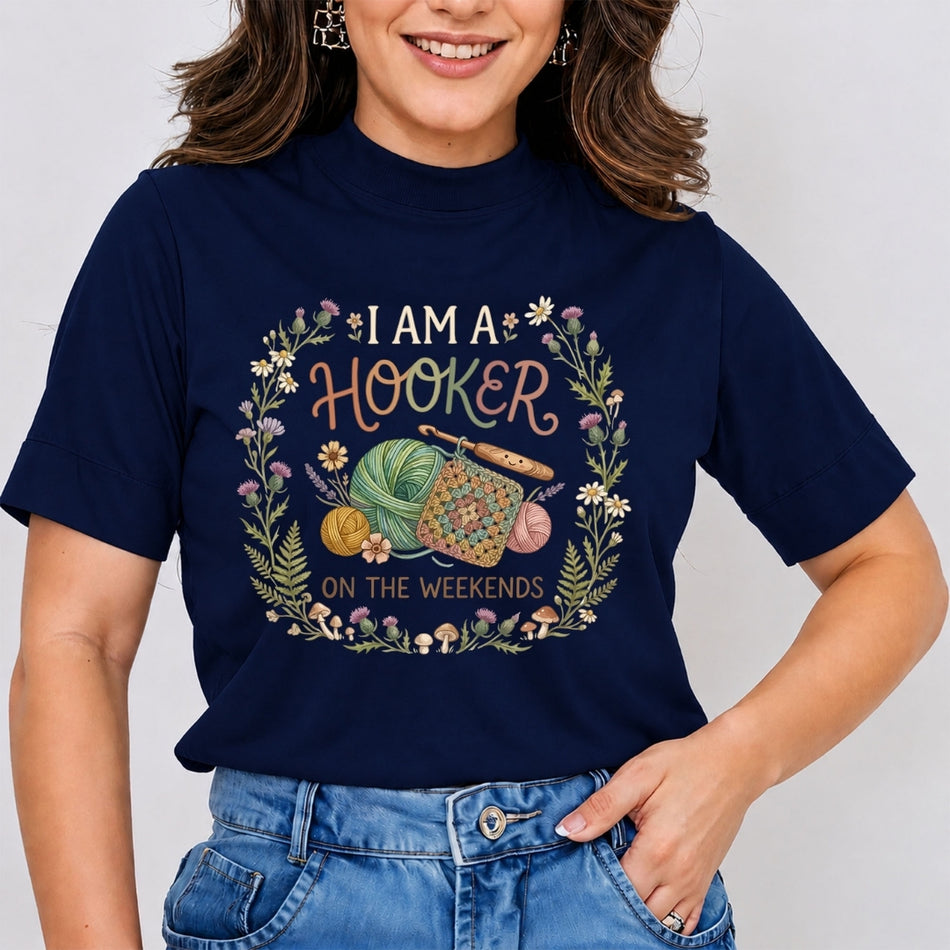 A woman wearing a navy blue graphic t-shirt with a floral wreath design and the text 'I AM A HOOKER ON THE WEEKENDS' along with knitting needles and yarn, paired with blue jeans.