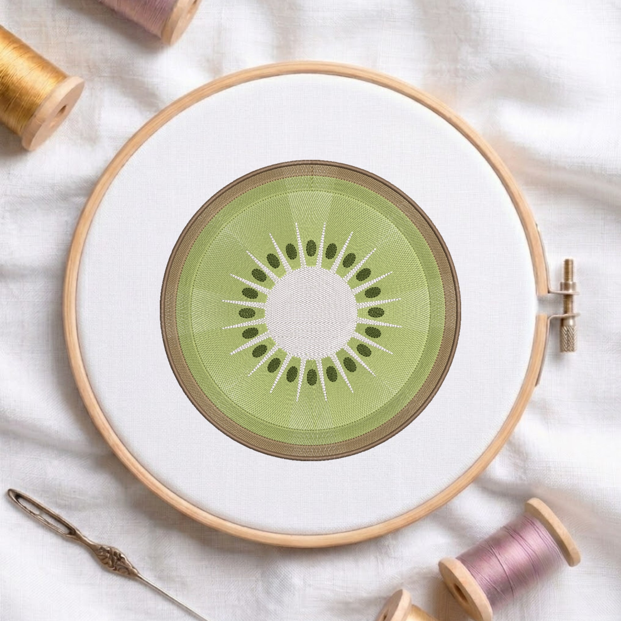 A circular embroidery hoop holding white fabric with a kiwi slice design stitched in the center. The hoop is photographed on a white cloth with spools of thread and a needle around it.