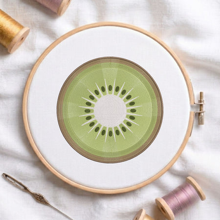 A circular embroidery hoop holding white fabric with a kiwi slice design stitched in the center. The hoop is photographed on a white cloth with spools of thread and a needle around it.