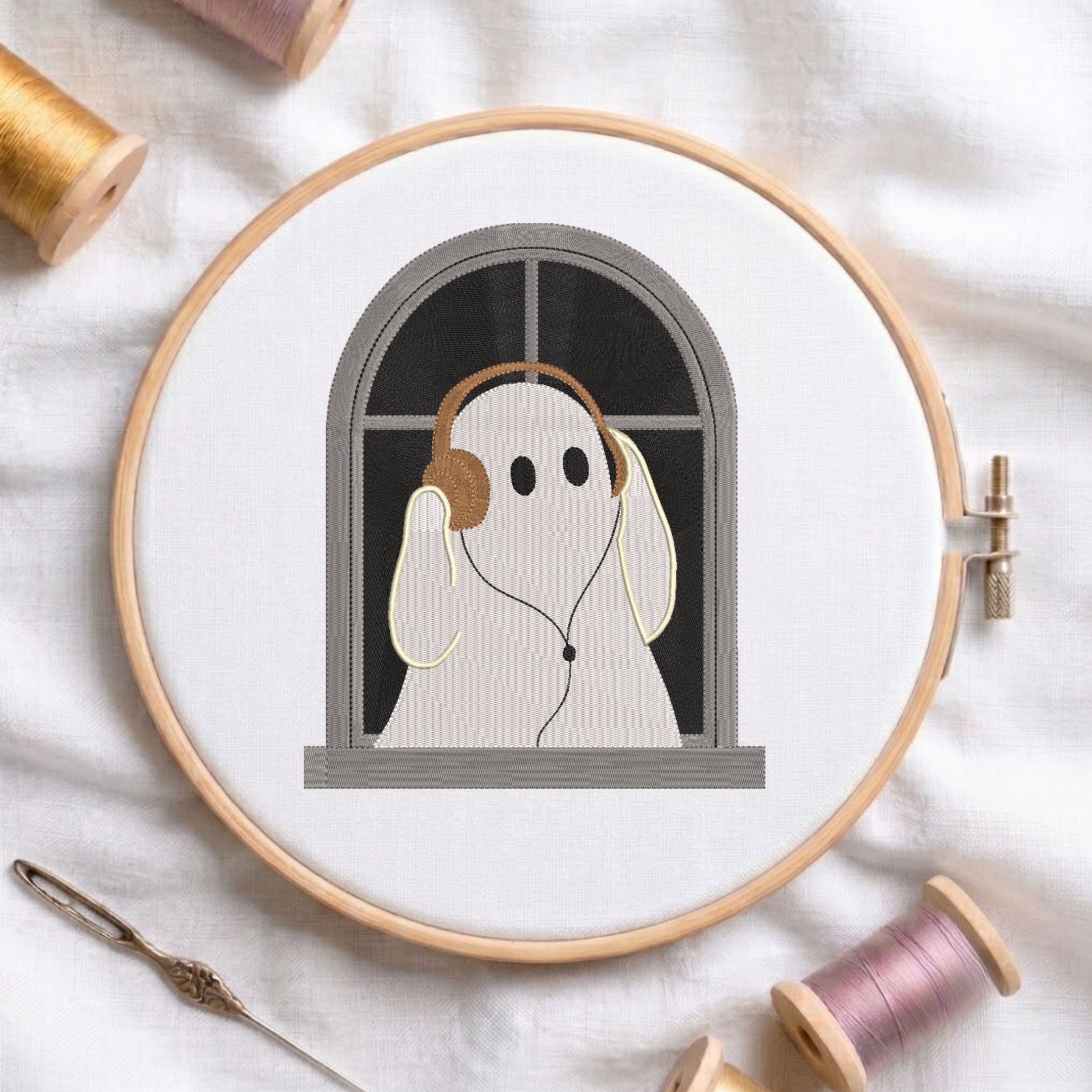 A circular embroidery hoop displays a stitched design of a ghost-like figure wearing brown headphones, framed by a light wood hoop on a white fabric background with spools of thread and a needle.