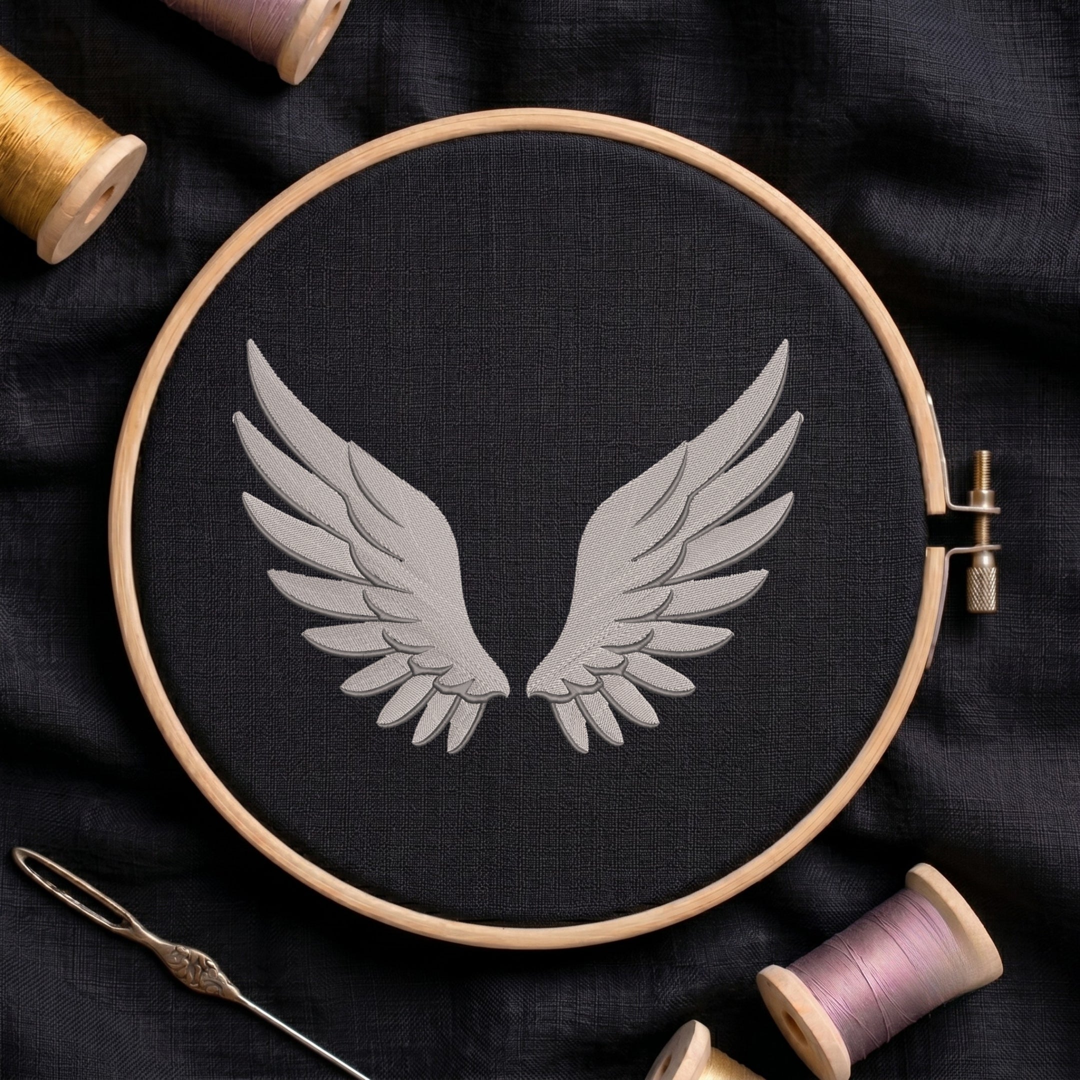 A circular embroidery hoop holding black fabric with a light gray embroidered pair of wings, surrounded by spools of thread and a needle on a dark fabric background.