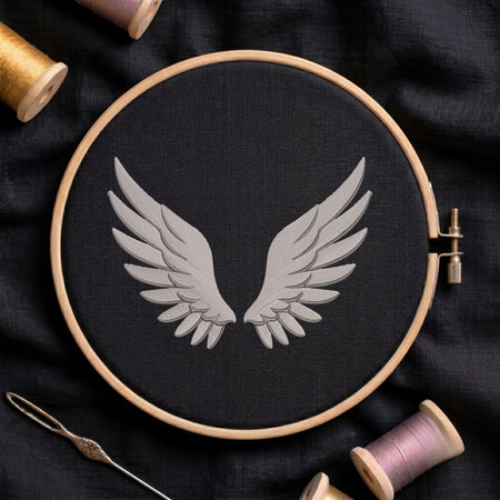 A circular embroidery hoop holding black fabric with a light gray embroidered pair of wings, surrounded by spools of thread and a needle on a dark fabric background.