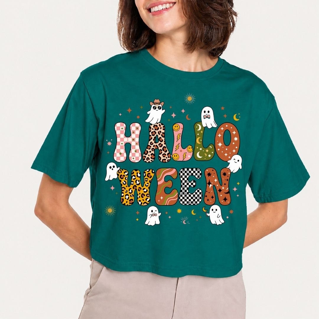 A woman wearing a teal green cropped t-shirt with a colorful 'HALLOWEEN' graphic featuring ghosts, stars, and patterned letters, paired with beige pants against a light gray background.