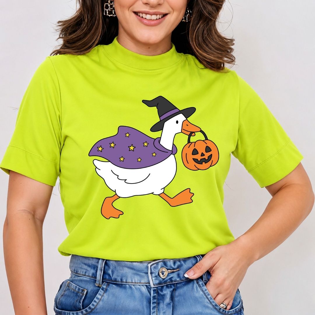 A woman wearing a bright lime green mock-neck t-shirt with a cartoon graphic of a white goose dressed as a witch in a purple cape and black hat, holding an orange jack-o'-lantern. She is paired with blue jeans against a plain light background.