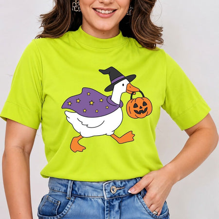 A woman wearing a bright lime green mock-neck t-shirt with a cartoon graphic of a white goose dressed as a witch in a purple cape and black hat, holding an orange jack-o'-lantern. She is paired with blue jeans against a plain light background.