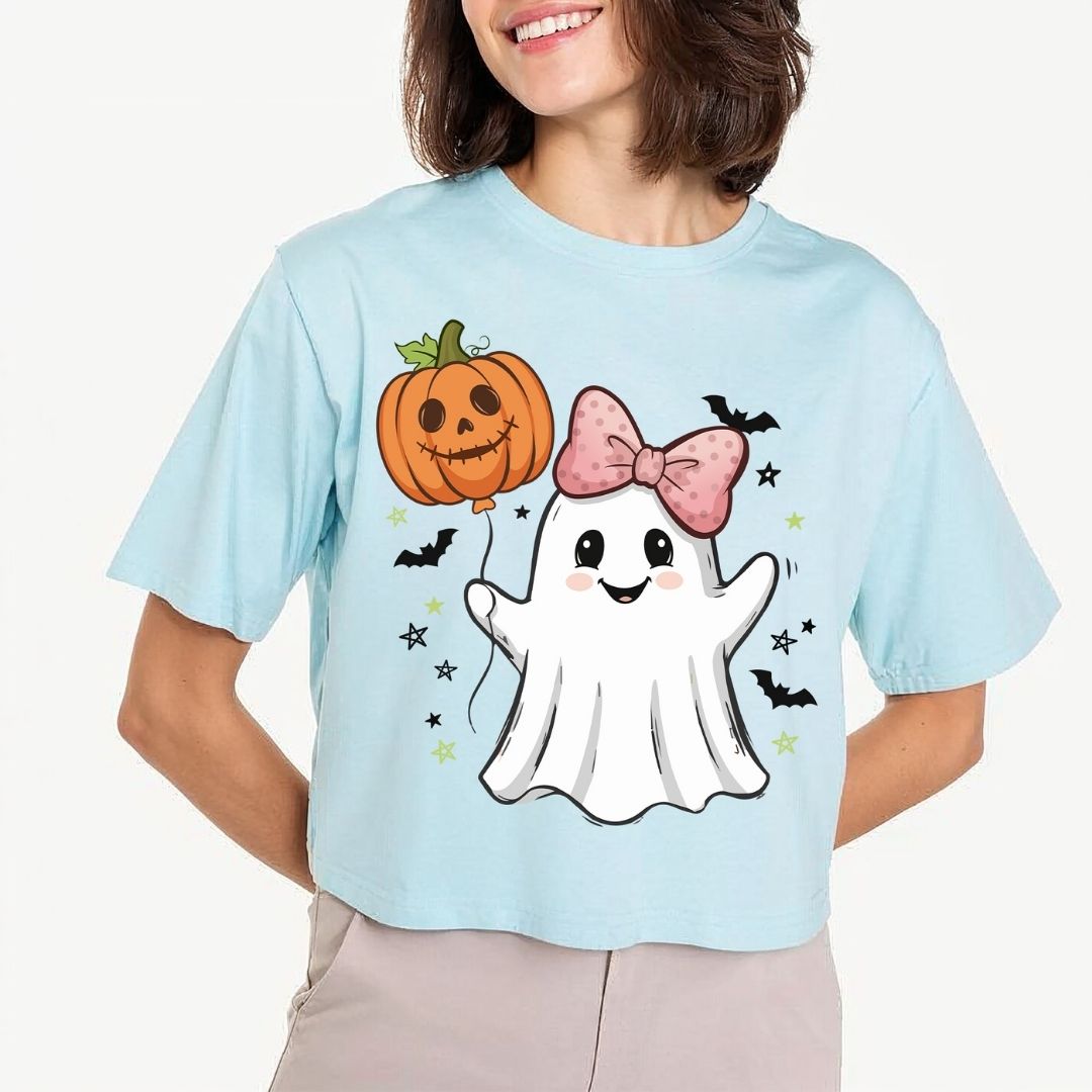 A woman wearing a light blue cropped t-shirt with a cute Halloween graphic of a white ghost holding an orange jack-o'-lantern balloon, surrounded by black bats and stars.
