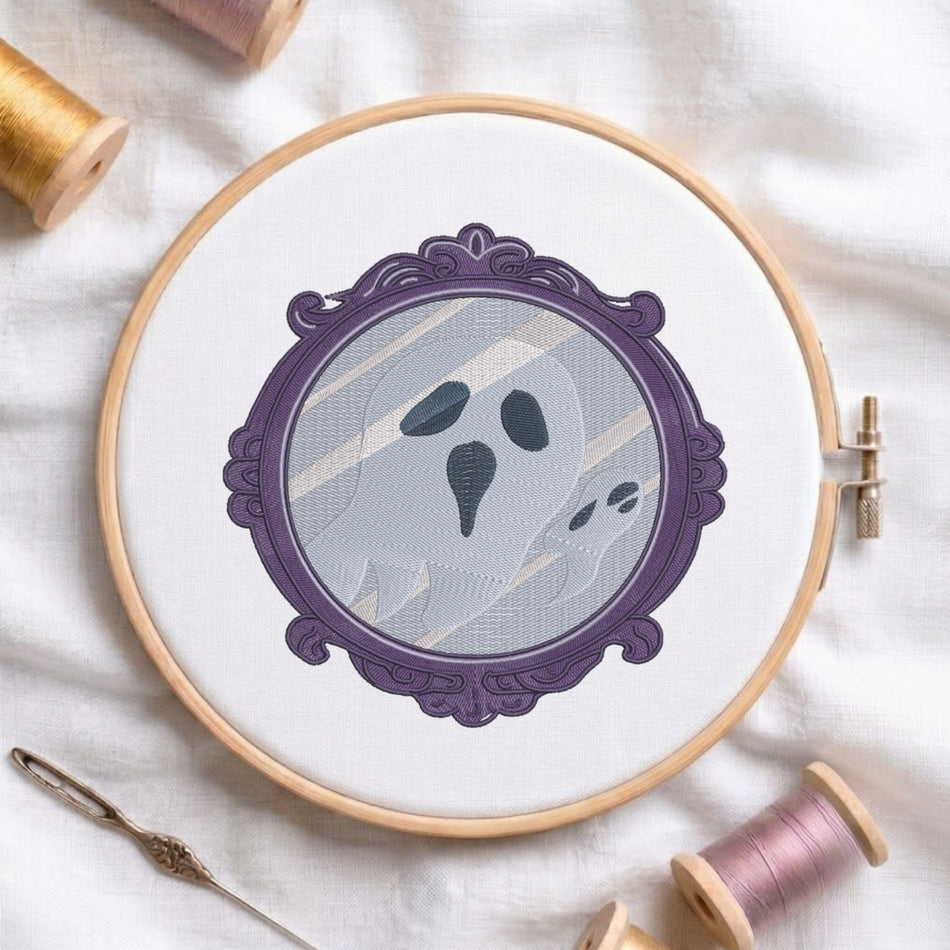 A circular embroidery hoop holding white fabric with a ghost design framed in purple, surrounded by spools of thread and a needle on a white cloth background.